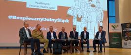 Na scenie odbywa się panel dyskusyjny. Siedmiu mężczyzn siedzi na krzesłach w rzędzie. Jeden z nich, w mundurze strażackim, trzyma mikrofon i mówi. Pozostali panowie są ubrani w garnitury. Za nimi na ekranie widnieje napis „Panel dyskusyjny” oraz hasło „#BezpiecznyDolnyŚląsk”.
