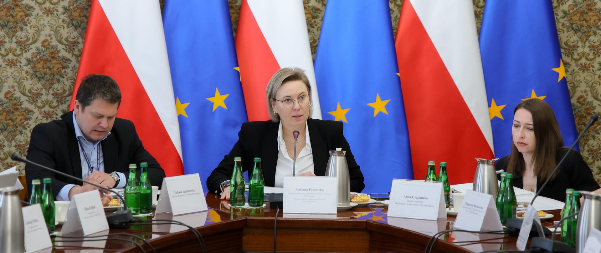 Zdjęcie sytuacyjne. Zbliżenie. Na zdjęciu, przy stole konferencyjnym, widoczni od lewej Pan Łukasz Jachimowicz, Dyrektor Departamentu Społeczeństwa Obywatelskiego w KPRM, Pani Adriana Porowska, Sekretarz Stanu w Kancelarii Prezesa Rady Ministrów, Wiceprzewodnicząca Komitetu do spraw Pożytku Publicznego i Pani Anna Czaplińska, Zastępca Dyrektora w Departamencie Społeczeństwa Obywatelskiego w KPRM. W tle stoją ustawione w rzędzie flagi Polski i Unii Europejskiej.