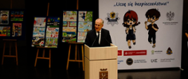 Mężczyzna przemawia przy mównicy za nimi na sztalugach ułożone są prace plastyczne oraz baner akcji Uczę się bezpieczeństwa" z logo Policji oraz podmiotów partnerów wspierających akcję. 