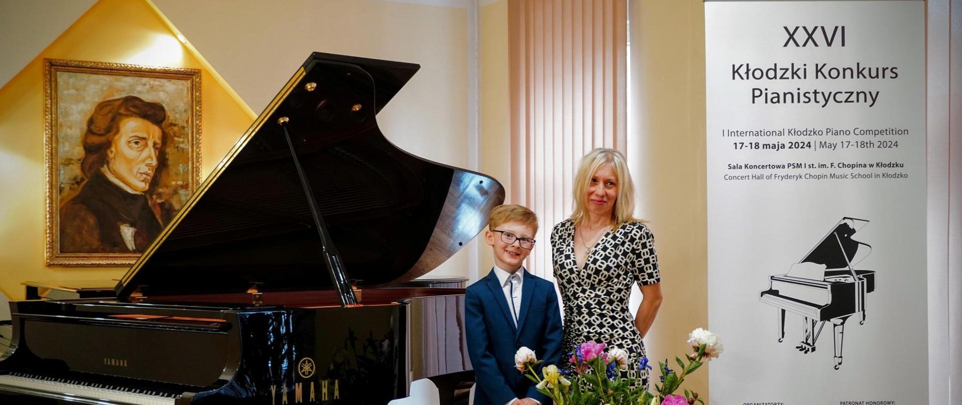 uczeń z nauczycielką na tle fortepianu, portretu Chopina, baner z napisem XXVI Kłodzki Konkurs Pianistyczny