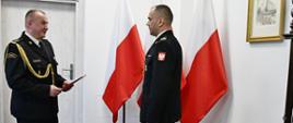 Strażak w mundurze wyjściowym ze sznurem trzyma czerwoną teczkę przed nim stoi strażak w mundurze wyjściowym obok stoją trzy flagi Polski są drzwi na ścianie wisi obraz oraz obok stoi zegar. 