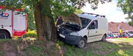 Pojazd osobowy VW Caddy koloru białego uderzył w przydrożne drzewo. W tle widoczne są 2 karetki pogotowia oraz tylna część wozu strażackiego.
