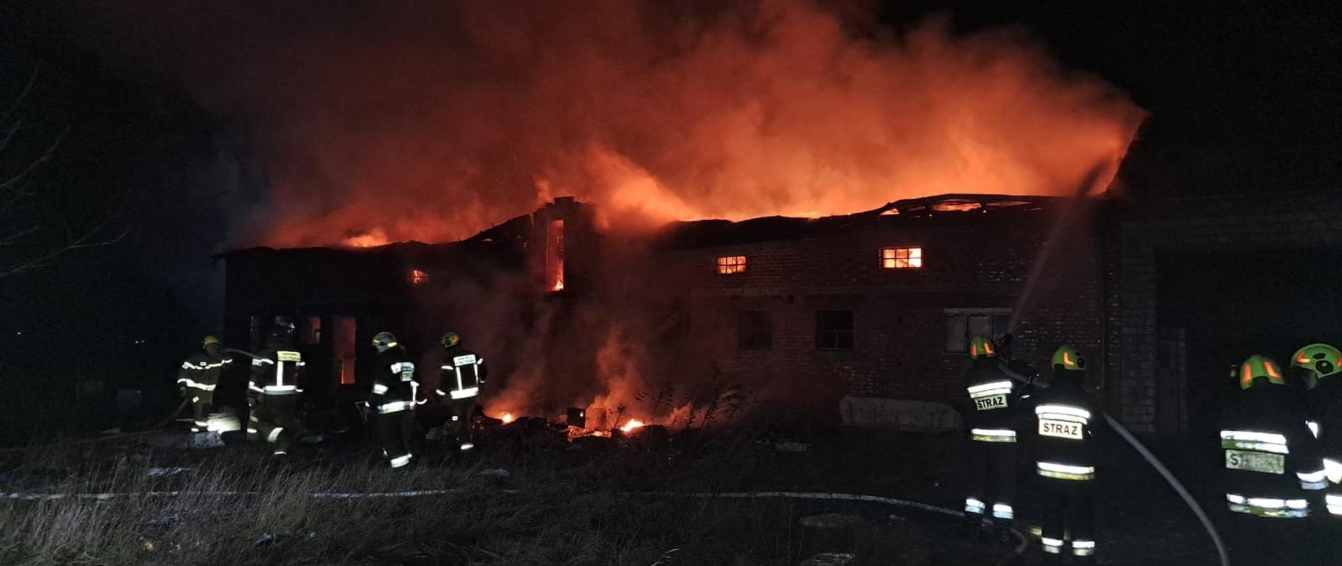 Na zdjęciu widać palący się budynek gospodarczy. Strażacy polewają go wodą. Przed budynkiem strażacy gaszą pożar elementów konstrukcji budynku, które spadły na zmienię. Są ubrani w piaskowe stroje i żółte hełmy. Budynek nie ma dachu i szyb w oknach. Jest ciemno.