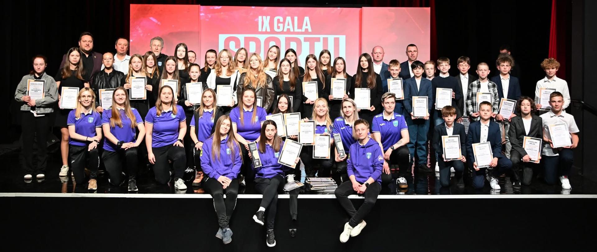 IX Gala Sportu w Biskupcu: na scenie wiele osób, organizatorzy, opiekunowie, dzieci i młodzież z dyplomami.