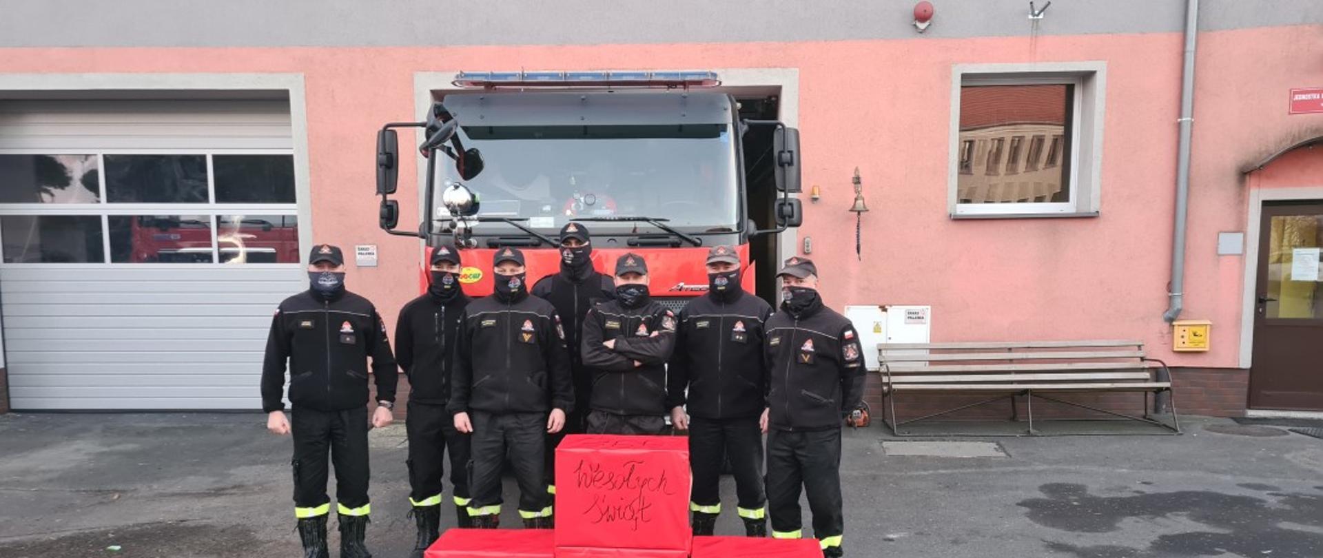 Obraz przedstawia strażaków Jednostki Ratowniczo-Gaśniczej w Jaworze w umundurowaniu koszarowym na tle samochodu pożarniczego i budynku komendy. Przed strażakami stoją cztery zapakowane w czerwony papier prezenty na których napisano wesołych świąt KP PSP Jawor.