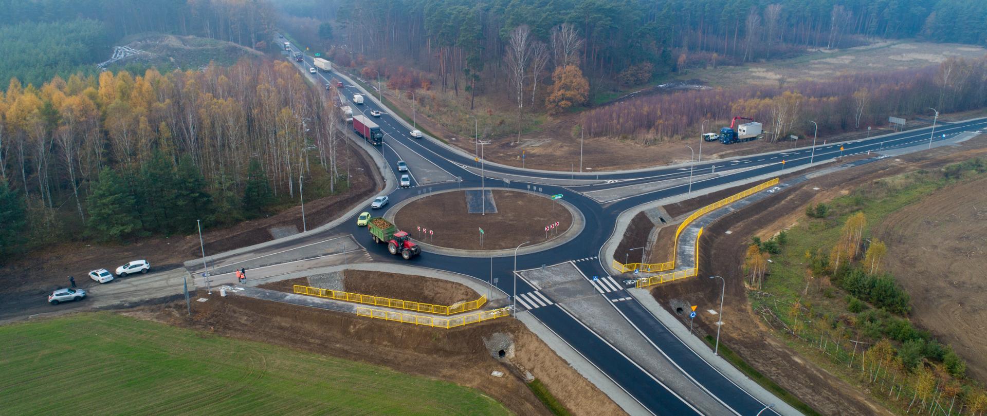 Zdjęcie z powietrza. Nowe czterowlotowe rondo na skrzyżowaniu dróg z przejściem dla pieszych i droga rowerową w obrębie skrzyżowania. W otoczeniu lasy i pola.