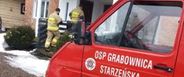 Kolorowe zdjęcie przedstawia na pierwszym planie, po prawej stronie, czerwony, oznakowany samochód lekki Ochotniczej Straży Pożarnej z napisem OSP Grabownica Starzeńska. Na środkowym planie widoczne dwie sylwetki umundurowanych w jasne ubrania strażaków z napisami na plecach STRAŻ, wchodzących do budynku mieszkalnego. Widoczna niewielka ilość śniegu.