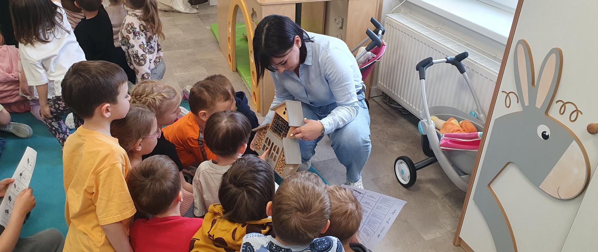 Inspektor w przedszkolu klęczy na podłodze i pokazuje dzieciom drewniany model domku. Wokół niej siedzi grupa małych dzieci, które z zainteresowaniem oglądają przedmiot. W tle widać salę zabaw z wyposażeniem dla dzieci, wózek dziecięcy oraz okno z kaloryferem. 