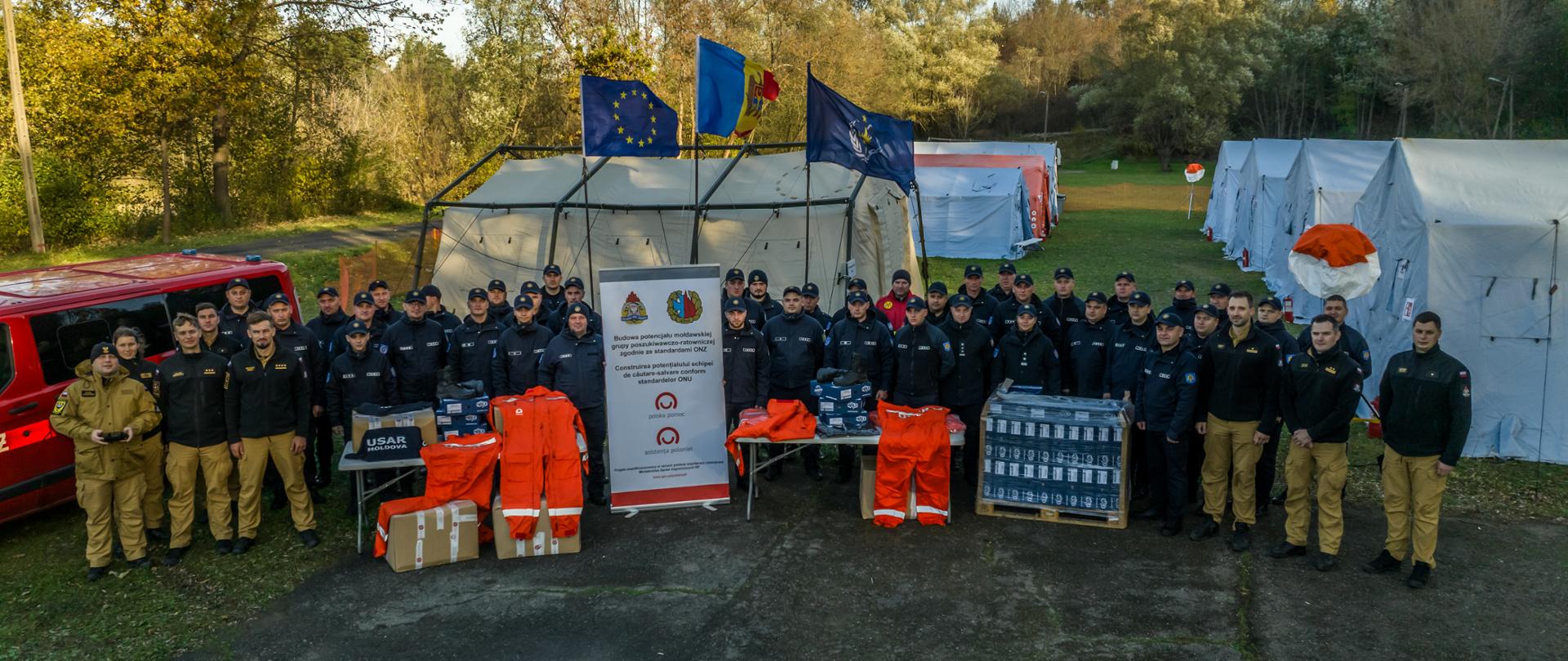 Zdjęcie grupowe polskich i mołdawskich ratowników na tle namiotów i sprzętu ratowniczego. Widoczne są flagi Unii Europejskiej, Mołdawii i Polski oraz baner programu „Polska Pomoc”.