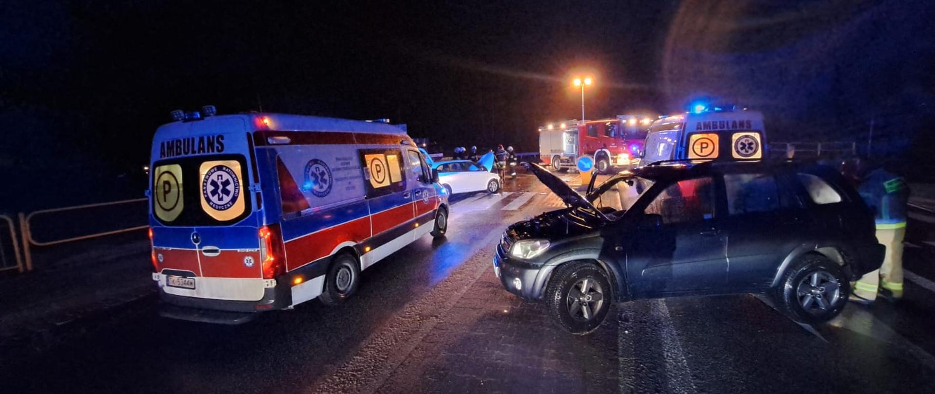 Zdjęcie przedstawia drogę wojewódzką gdzie doszło do zderzenia dwóch pojazdów osobowych. Jeden z nich stoi po prawej stronie, zaś po prawej widać ambulans Zespołu Ratownictwa Medycznego. W głębi stoją inne wozy służb ratowniczych