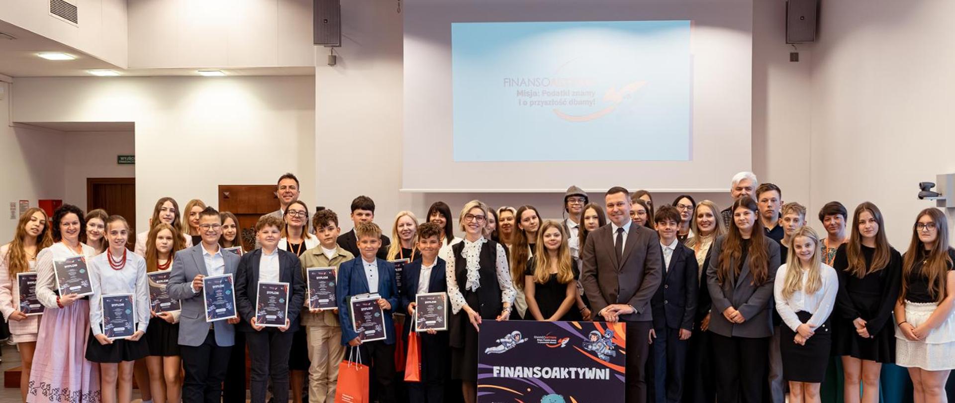 Grupa osób stoi w sali konferencyjnej, część trzyma dyplomy, a na środku widoczny jest plakat „FINANSOAKTYWNI” przed ekranem projekcyjnym