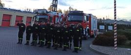 Zdjęcie przedstawia strażaków z JRG w Pruszczu Gdańskim stojących w dwuszeregu podczas uroczystej zmiany służby na tle dwóch samochodów strażackich, obok masztu flagowego z podniesioną flagą państwową.
