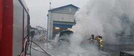 Zdjęcie przedstawia pożar samochodu osobowego przed budynkiem warsztatu samochodowego. Silne zadymienie. Strażacy podają wodę oraz pianę gaśniczą na palący się pojazd. Strażacy pracują w aparatach ochrony układu oddechowego. 