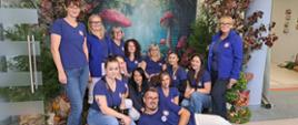 Zdjęcie przedstawia grupę pracowników PSSE w Jeleniej Górze ubranych w niebieskie koszulki z logo Państwowej Inspekcji Sanitarnej na tle lasu z czerwonymi muchomorami wielkości dorosłego człowieka.