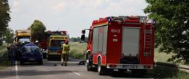 Zdjęcie przedstawia samochody służb ratowniczych i pojazdy biorące udział w zdarzeniu stojące na jezdni w miejscu, gdzie doszło do zdarzenia drogowego.