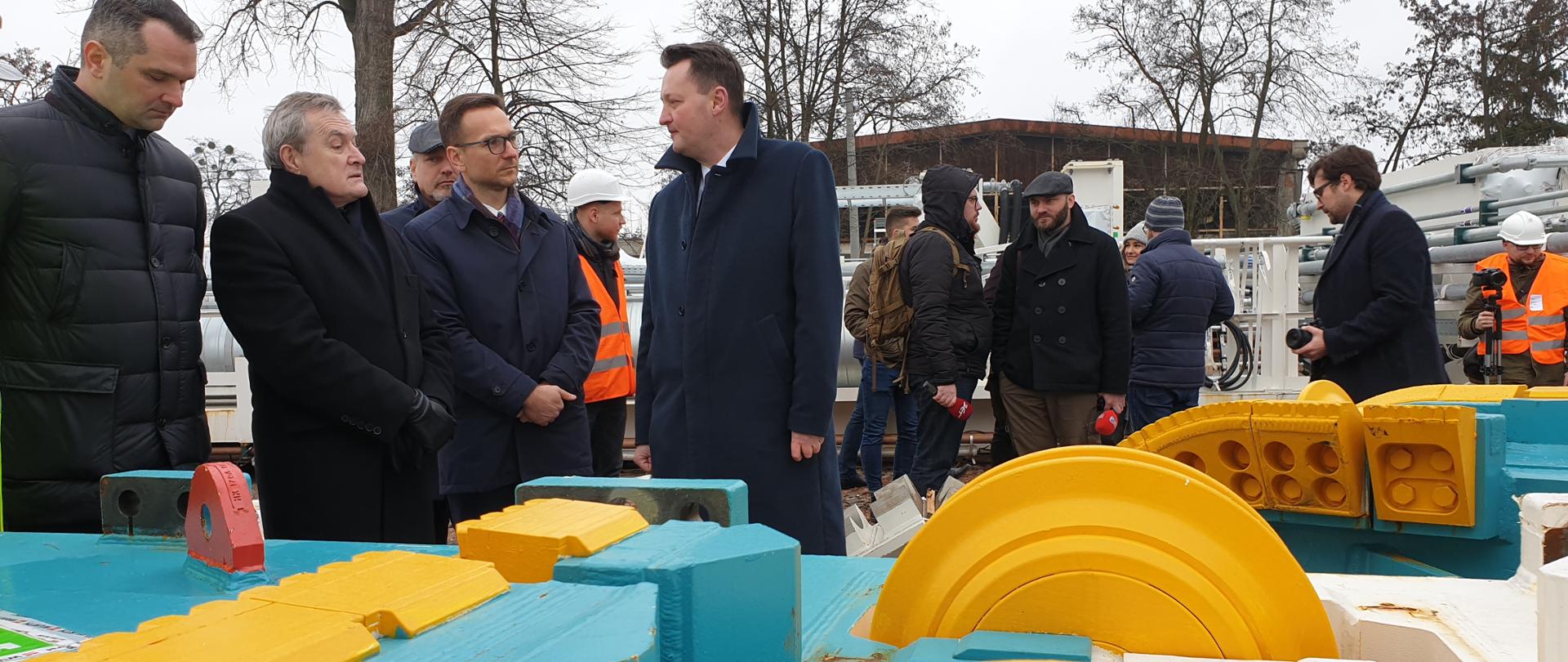 Na zdjęciu stoi kilkoro osób, w środku wicepremier Piotr Glińskie i wiceminister Waldemar Buda, w przodu kolorowe elementy tarczy, która będzie drążyła tunel pod Łodzią.