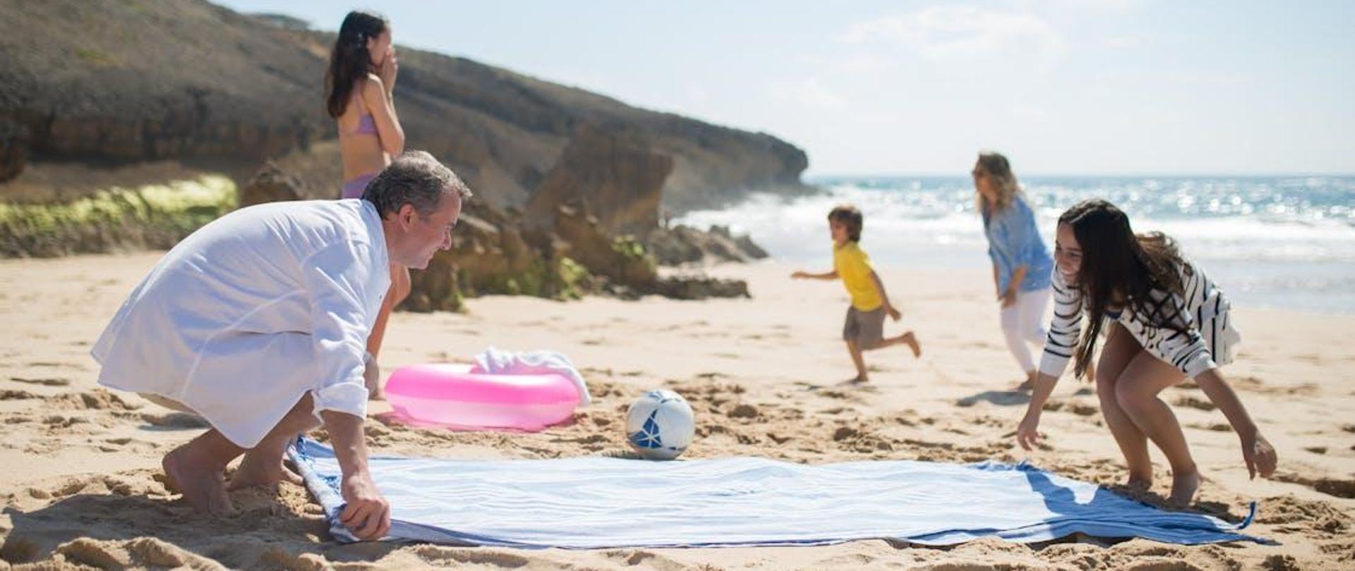 Rodzina wypoczywająca nad morzem. Doriśli rozkłądają na pisaku koc. Dzieci grają w piłkę. 