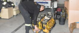 Sprzęt pożarniczy dla ukraińskich strażaków. Komenda Powiatowa PSP w Rawiczu. Strażacy przygotowują do transportu zebrany sprzęt. Jeden z nich przeciera szmatka buty specjalne. W tle przyczepka, agregat prądotwórczy i meble.