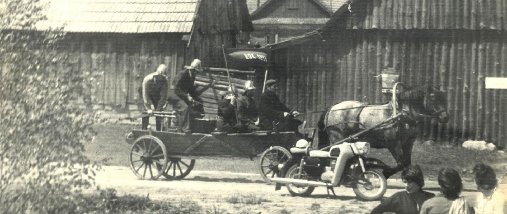 W zabudowie wiejskiej, drogą przejeżdża strażacki wóz konny. Obok motocykl i gapie.