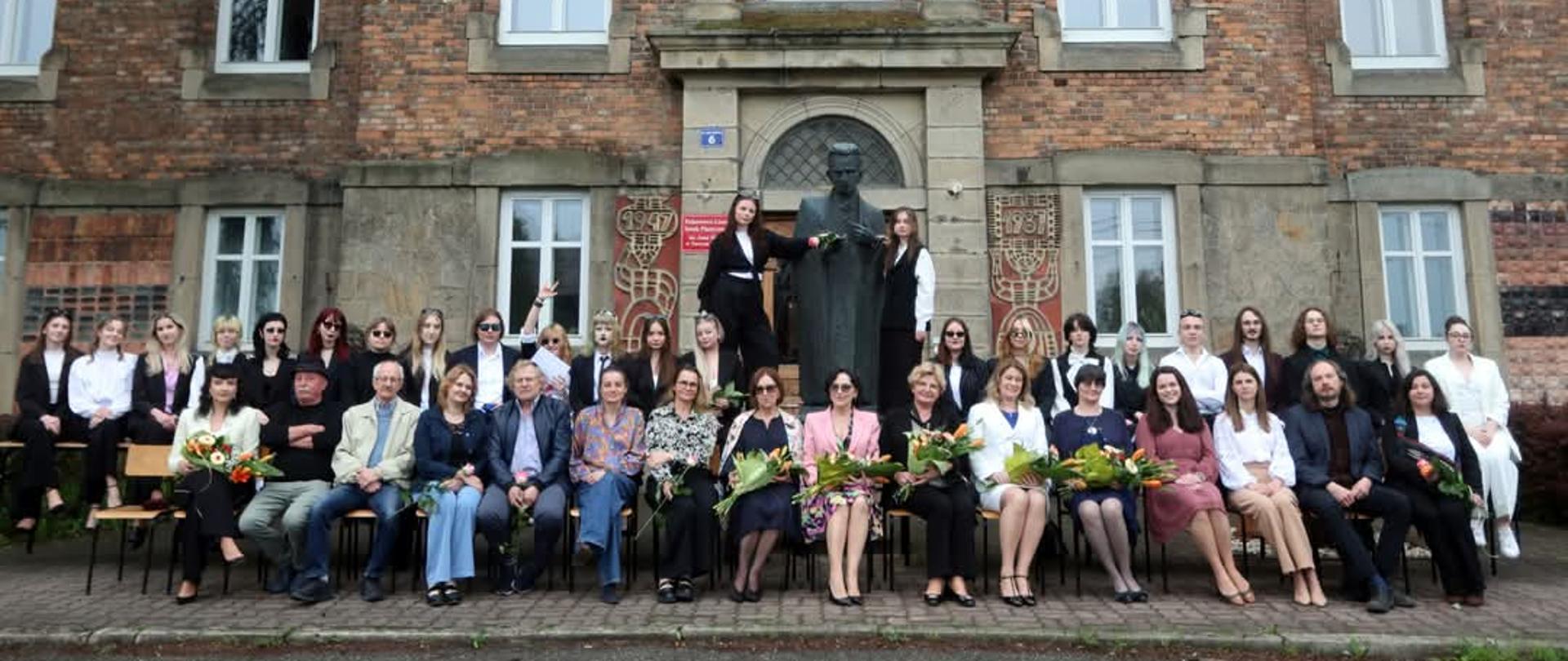 Zdjęcie ma charakter oficjalny. Grupa około 40 osób, absolwenci szkoły oraz grono pedagogiczne pozuje do wspólnego, pamiątkowego zdjęcia przed zabytkowym gmachem PLSP. W centrum znajduje się pomnik Stanisława Fischera, założyciela szkoły. Pani dyrektor, panie wicedyrektor, pani kierownik internatu oraz wychowawczynie klas trzymają bukiety kwiatów. 