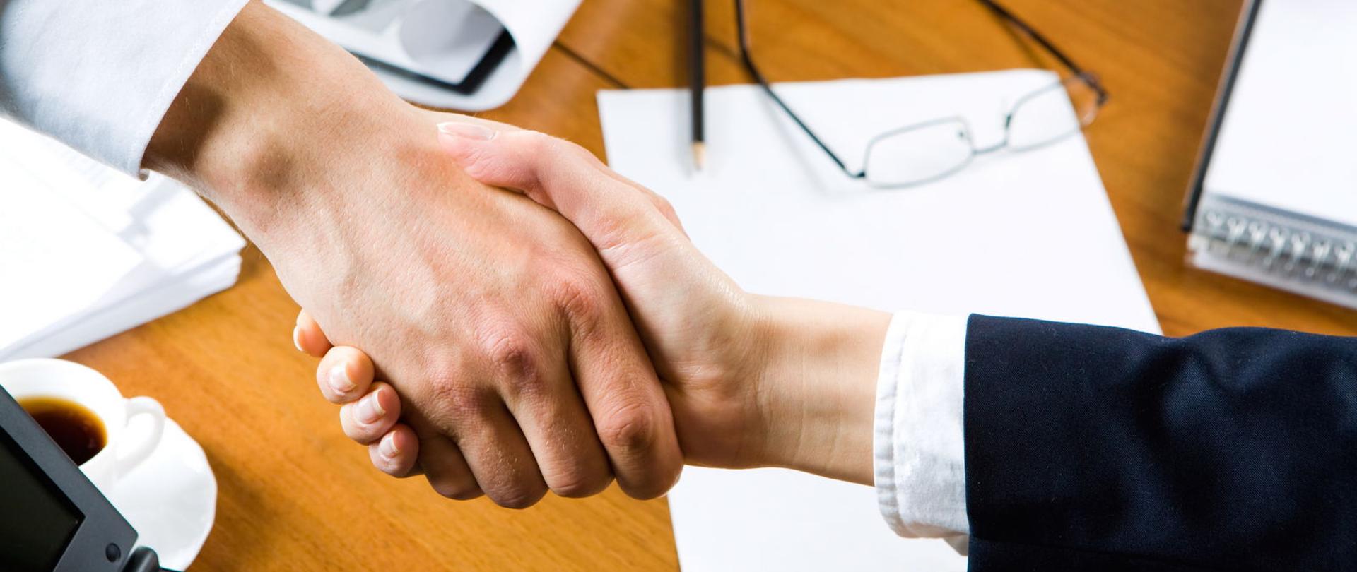 Uścisk dłoni, w tle stół z dokumentami i okulary.