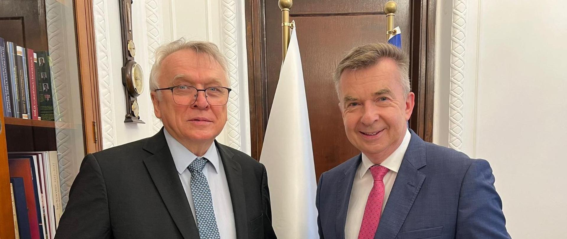 W gabinecie stoi minister Wieczorek i mężczyzna w czarnym garniturze, za nimi z boku przeszklona szafa z książkami.