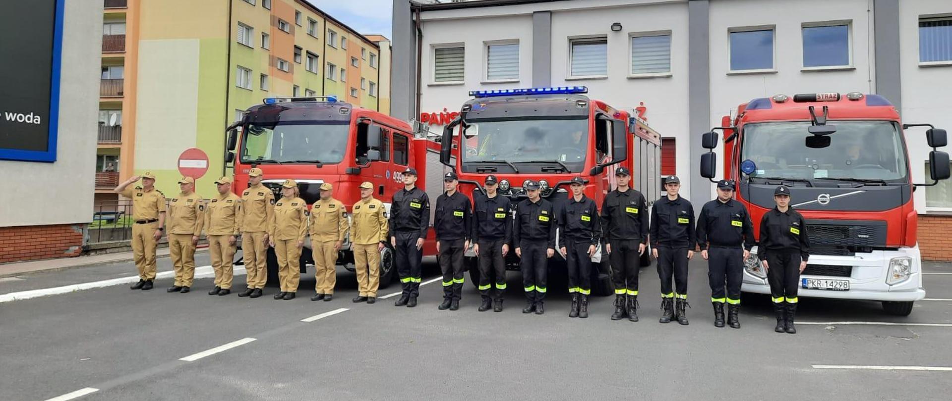 Strażacy stoją przed samochodami pożarniczymi, w szeregu, dowódca z prawej strony pododdziału salutuje. W tle remiza.