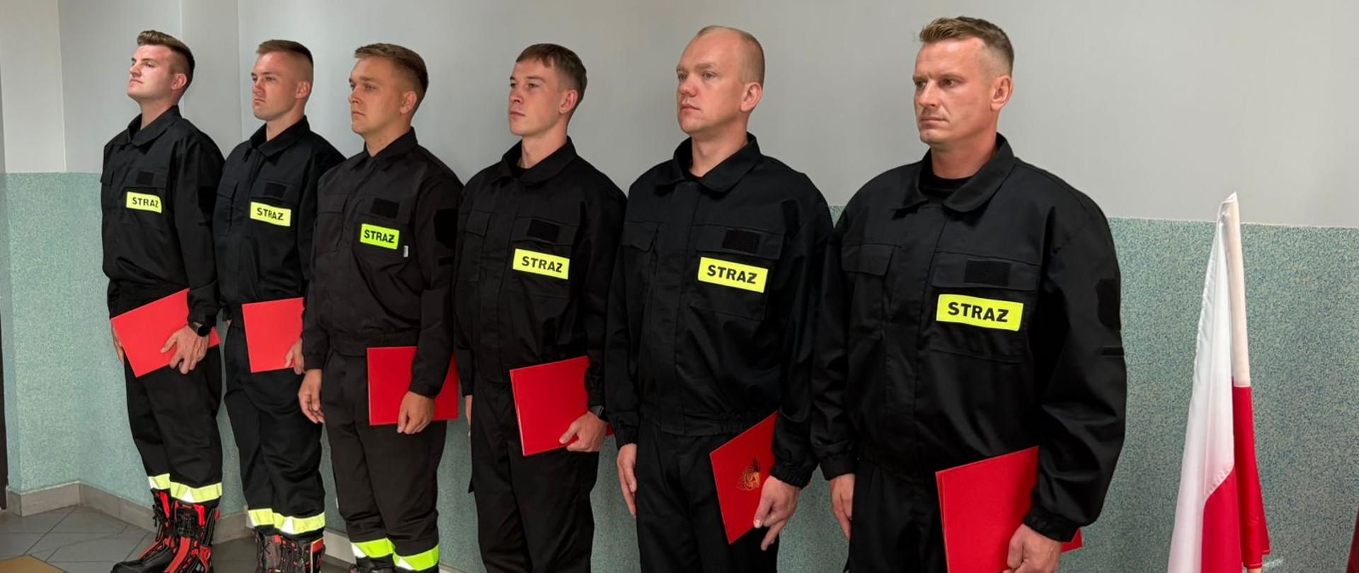 Strażacy stoją w szeregu po zakończonym ślubowaniu. W rękach trzymają teczki – akty ślubowania.
