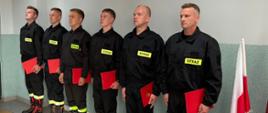 Strażacy stoją w szeregu po zakończonym ślubowaniu. W rękach trzymają teczki – akty ślubowania.