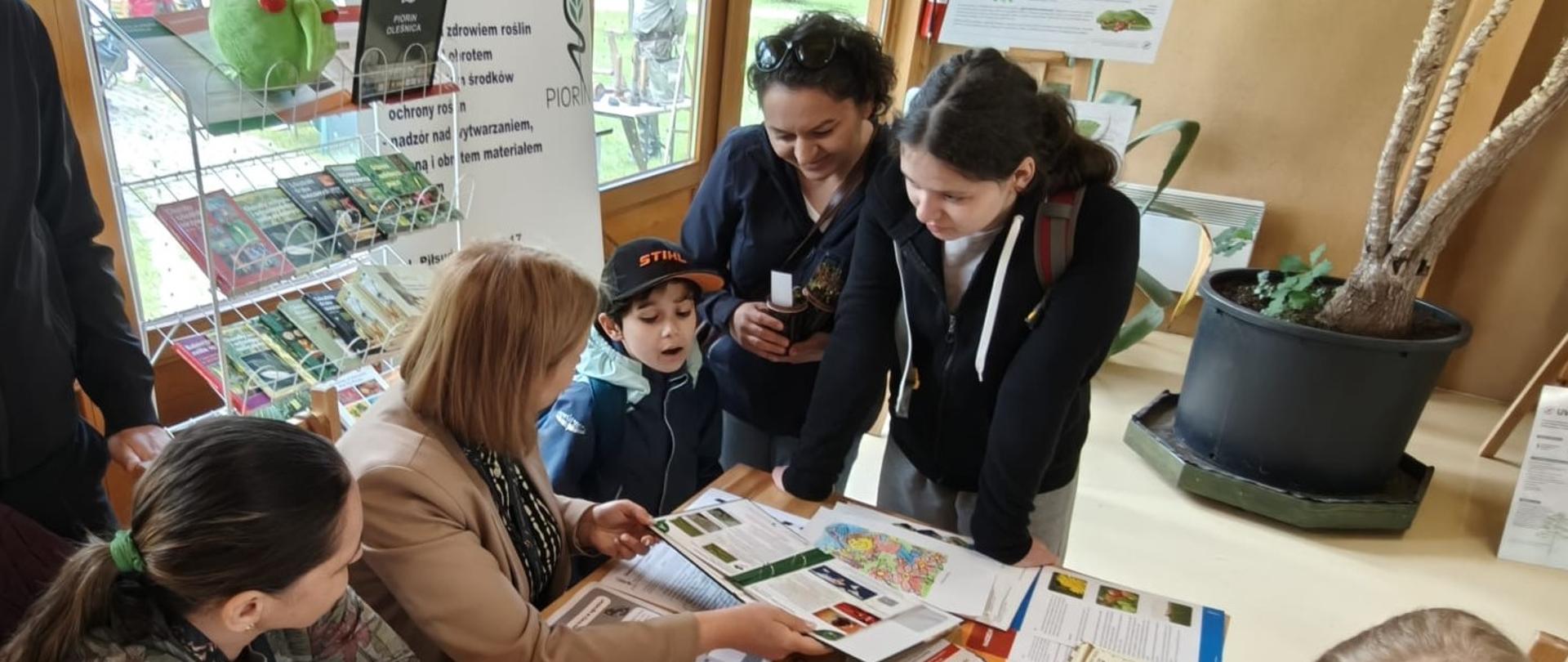 Przy stole na którym leżą rozłożone kolorowanki oraz materiały informacyjne inspekcji siedzi trójka dzieci, dwie dziewczynki kolorują obrazki. Obok panie inspektor z Oddziału oraz inni uczestnicy spotkania.