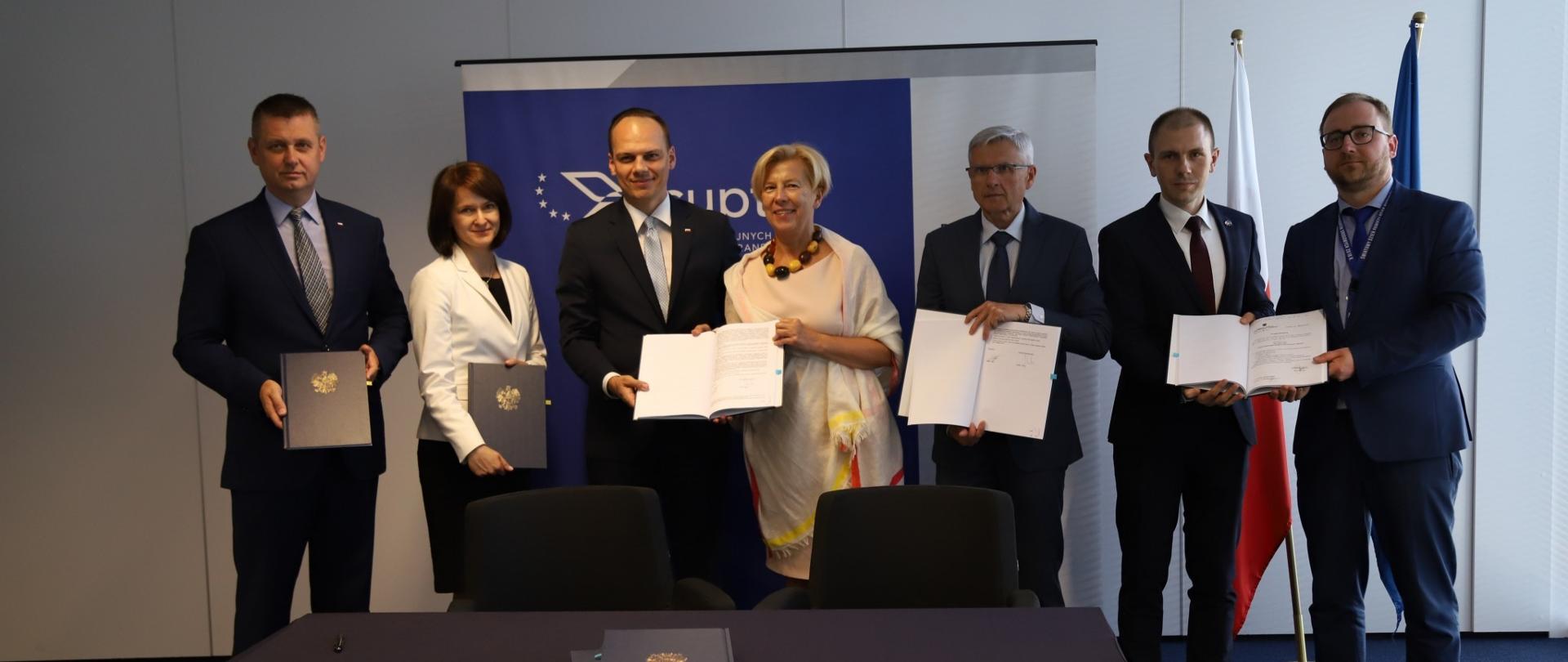 Na zdjęciu stoi 7 osób, w rękach trzymają podpisane umowy. Za nimi stoi rozstawiana ścianka z napisem "CUPT", flaga Polski i Unii Europejskiej. 