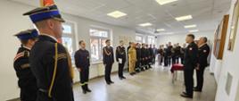 Na zdjęciu w mundurach galowych po prawej komendant powiatowy wraz z zastępcą po środku poczet sztandarowy ze sztandarem. Po lewej strażacy ubrani w czarne i piaskowe ubrania koszarowe oraz ubrania galowe a także pracownicy cywilni komendy uczestniczący w uroczystość ślubowania na sali konferencyjnej.