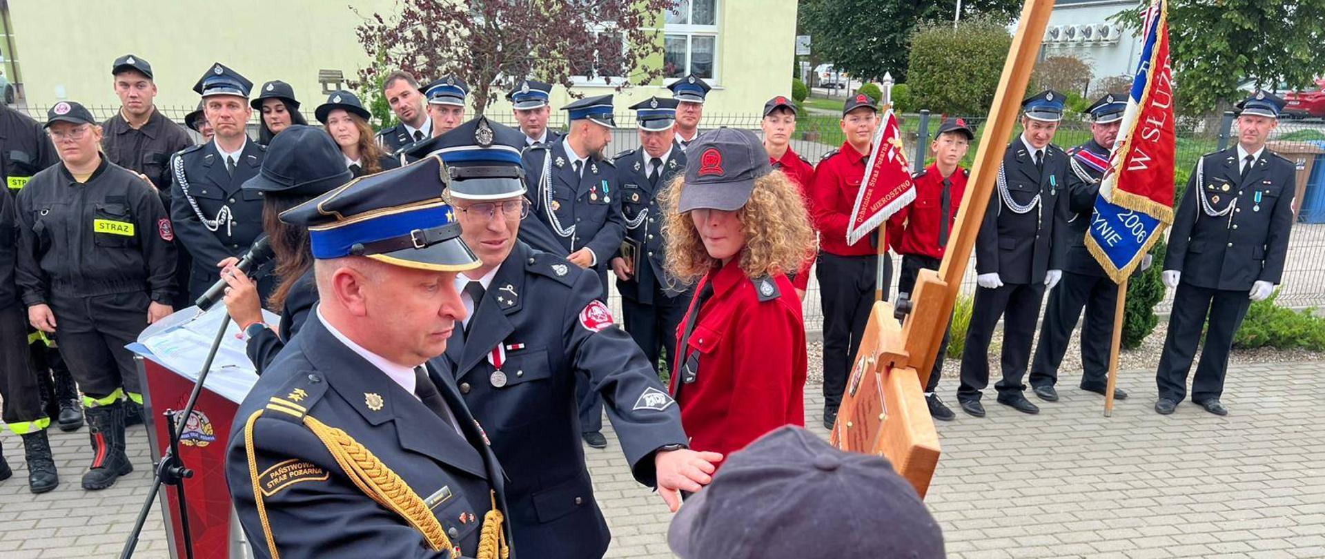 Komendant Powiatowy PSP w Pucku przybija symboliczną tabliczkę z okazji 20-lecia Młodzieżowej Drużyny Pożarniczej w Mieroszynie. Komendant jest ubrany w mundur galowy. 