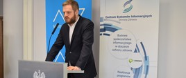 Wiceminister zdrowia Janusz Cieszyński na konferencji prasowej w Centrum Systemów Informacyjnych Ochrony Zdrowia. Wiceminister stoi za mównicą Ministerstwa Zdrowia, za jego plecami roll-upy resortu oraz CSIOZ.