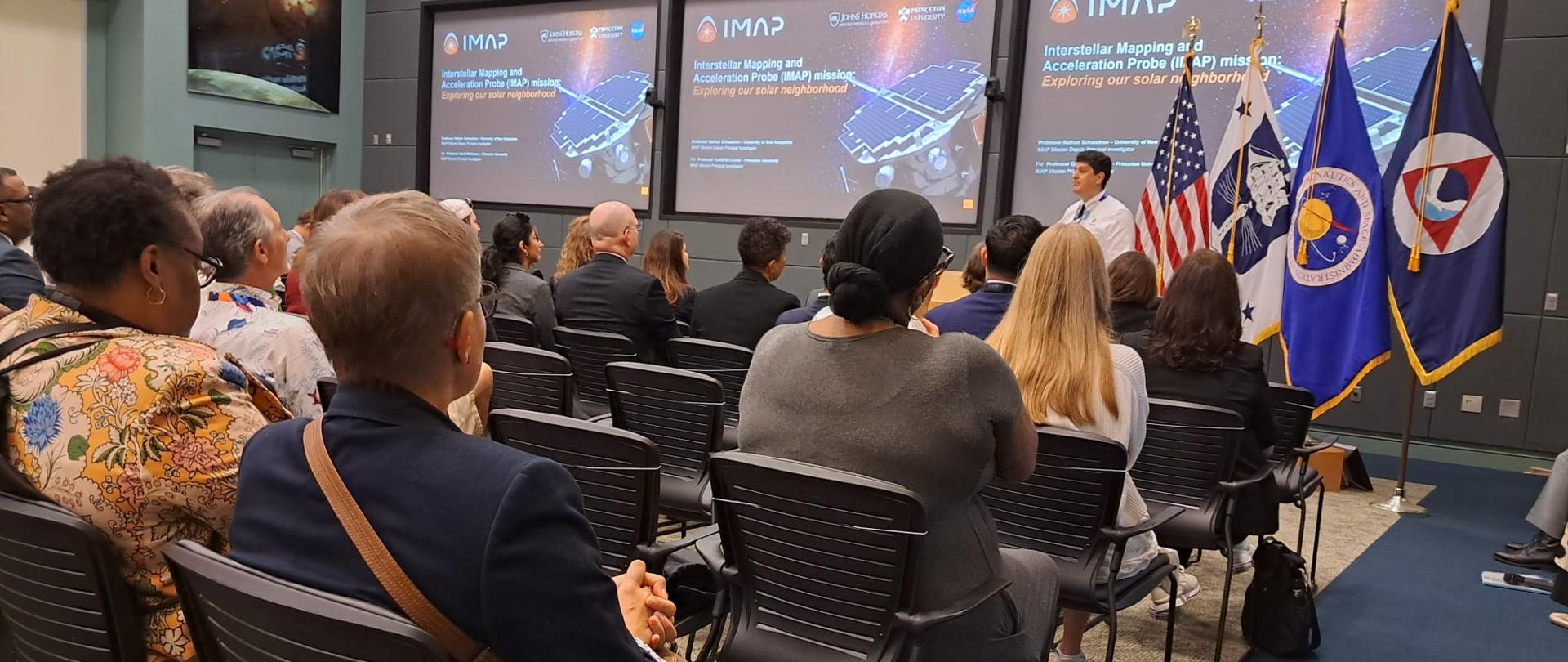 Zdjęcie od tyłu na salę, w rzędach krzeseł siedzą ludzie, przed nimi mężczyzna w białej koszuli mówi, za nim ściana na której wiszą 3 wielkie monitory.