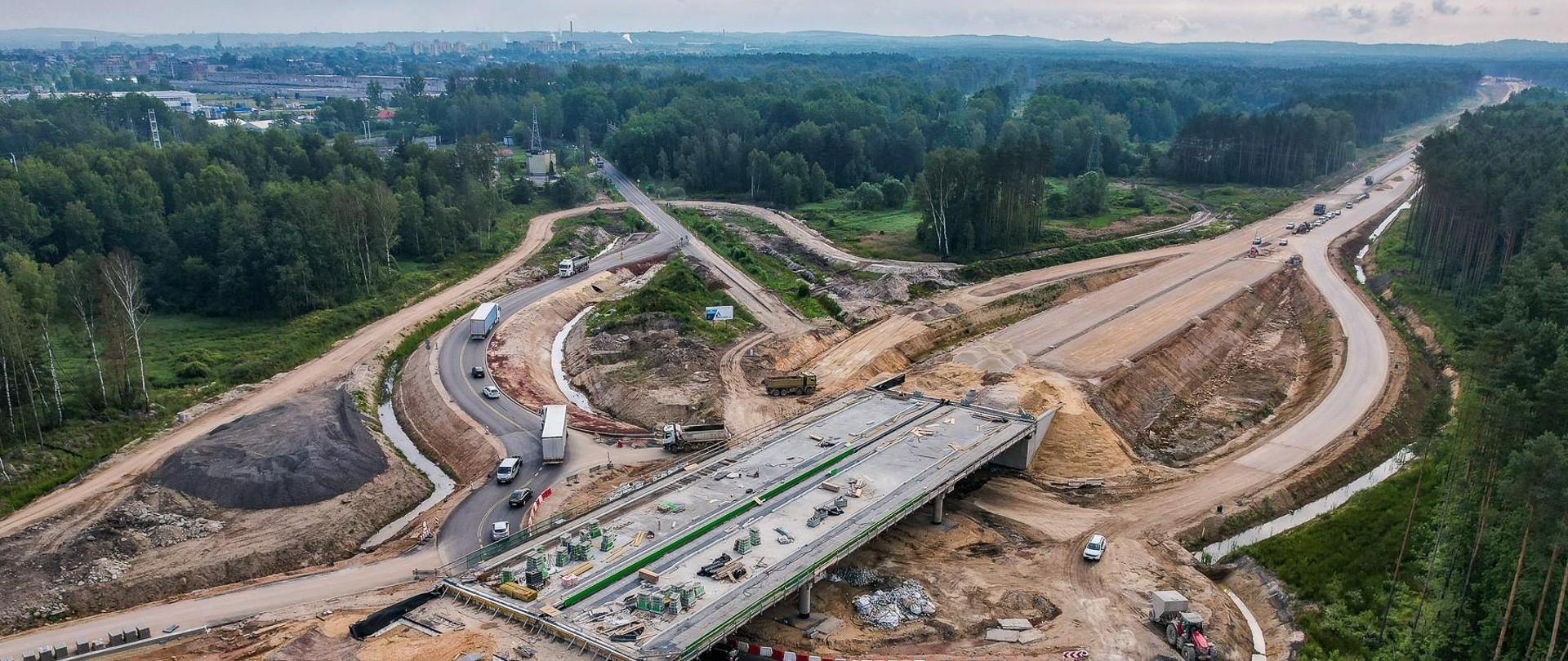 Zdjęcie przedstawia lotniczy widok na duży plac budowy drogi. Widoczny jest częściowo ukończony wiadukt, a pod nim przebiega istniejąca droga. Na miejscu widoczne są maszyny i pojazdy budowlane Otaczający krajobraz obejmuje fragmenty obszarów zalesionych, a w oddali znajduje się zabudowany obszar miasta.