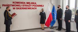 Akt ślubowania nowego funkcjonariusza. Osoby na zdjęciu ubrane w umundurowanie wyjściowe, uroczystość ma miejsce w sali konferencyjnej komendy. Osoba ślubująca ma podniesioną prawą rękę oraz dwa palce. Akt ślubowania odczytuje komendant miejski.