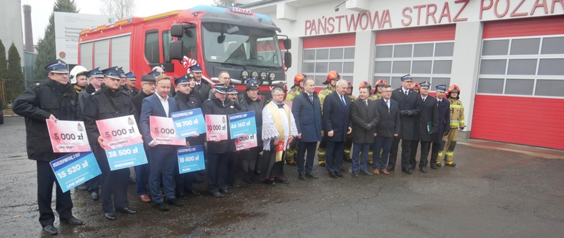 Fotografia przedstawia grupę strażaków ochotników, strażaków zawodowych oraz oficjeli rządowych i kościelnych pozujących na tle pojazdu pożarniczego. Na dalszym planie widoczny budynek strażnicy.