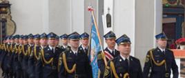 Kolumna strażaków w mundurach galowych, maszerujących w szyku przez wnętrze zabytkowego kościoła. Jeden z nich niesie niebieski sztandar z napisem „PSP w Gdańsku”. W tle widoczne są białe filary i elementy sakralne.