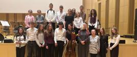 Na scenie oraz pod scenę w sali koncertowej Filharmonii Świętokrzyskiej pozują do zdjęcia uczniowie z nauczycielami oraz pośrodku wiolonczelistka z Filharmonii która jest jednocześnie nauczycielką. W tle widoczne są pulpity oraz krzesełka muzyków.
