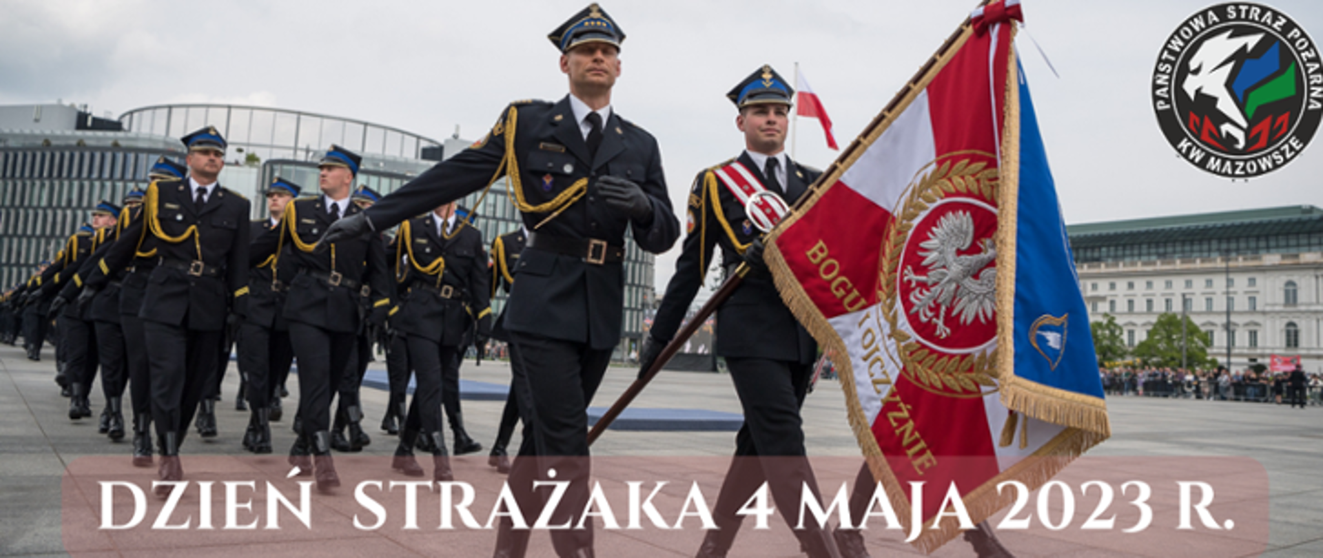 Zdjęcie wykonane na w Warszawie na Placu Marszałka Józefa Piłsudskiego. Na zdjęciu widać uroczystą defiladę pododdziałów Państwowej Straży Pożarnej z okazji obchodów Dnia Strażaka. Na pierwszym planie widać poczet sztandarowy. Poczet tworzy trzech funkcjonariuszy ubranych w mundury galowe. Środkowy z nich w rękach trzyma sztandar. Dodatkowo jego mundur przepasany jest biało – czerwoną wstęgą. Za pocztem sztandarowym maszerują w czwórkach pozostali funkcjonariusze również ubrani w mundury galowe. W oddali widać zabudowania okalające plac, na którym odbywa się uroczystość. 