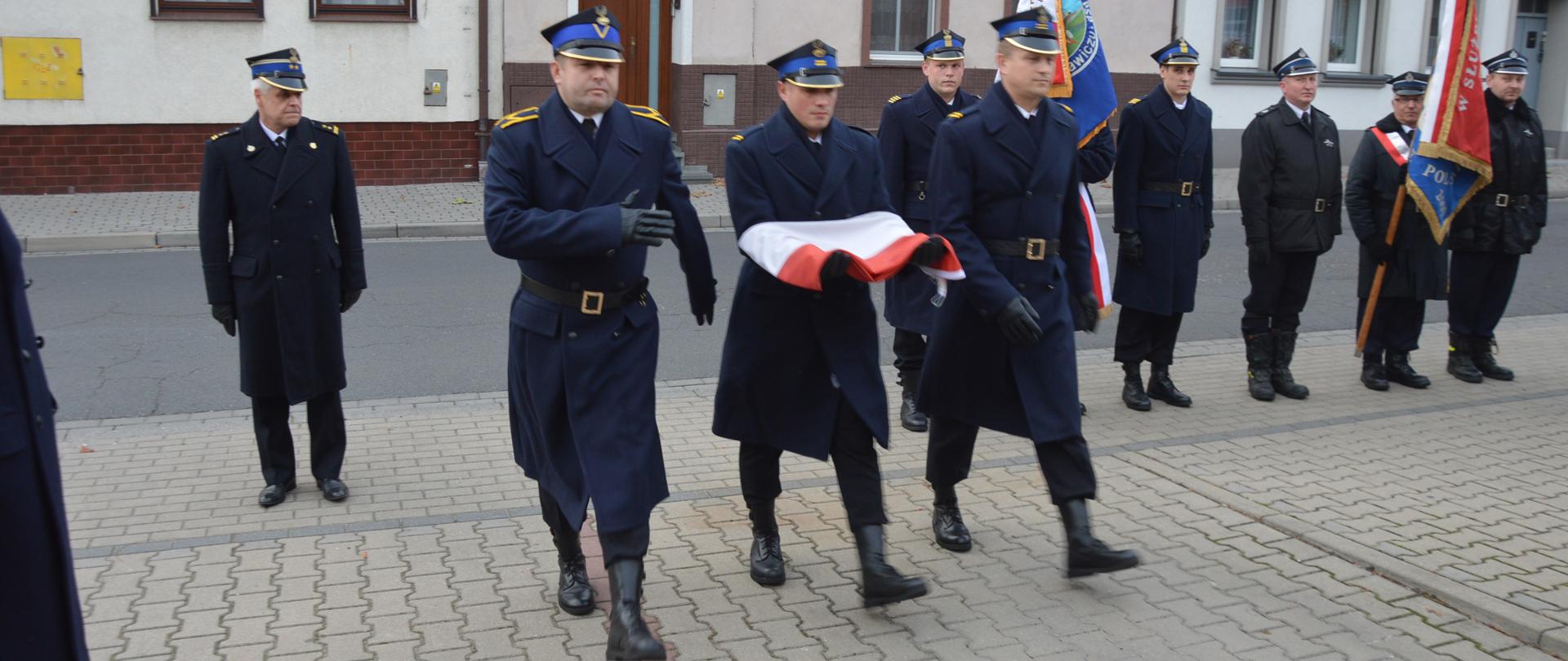 Narodowe Święto Niepodległości. Plac przed Komendą Powiatową PSP w Rawiczu. W kierunku masztu maszeruje poczet flagowy. Strażak w środku niesie rękach biało-czerwoną flagę. W tle budynki mieszkalne i poczty sztandarowe,