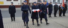 Narodowe Święto Niepodległości. Plac przed Komendą Powiatową PSP w Rawiczu. W kierunku masztu maszeruje poczet flagowy. Strażak w środku niesie rękach biało-czerwoną flagę. W tle budynki mieszkalne i poczty sztandarowe,