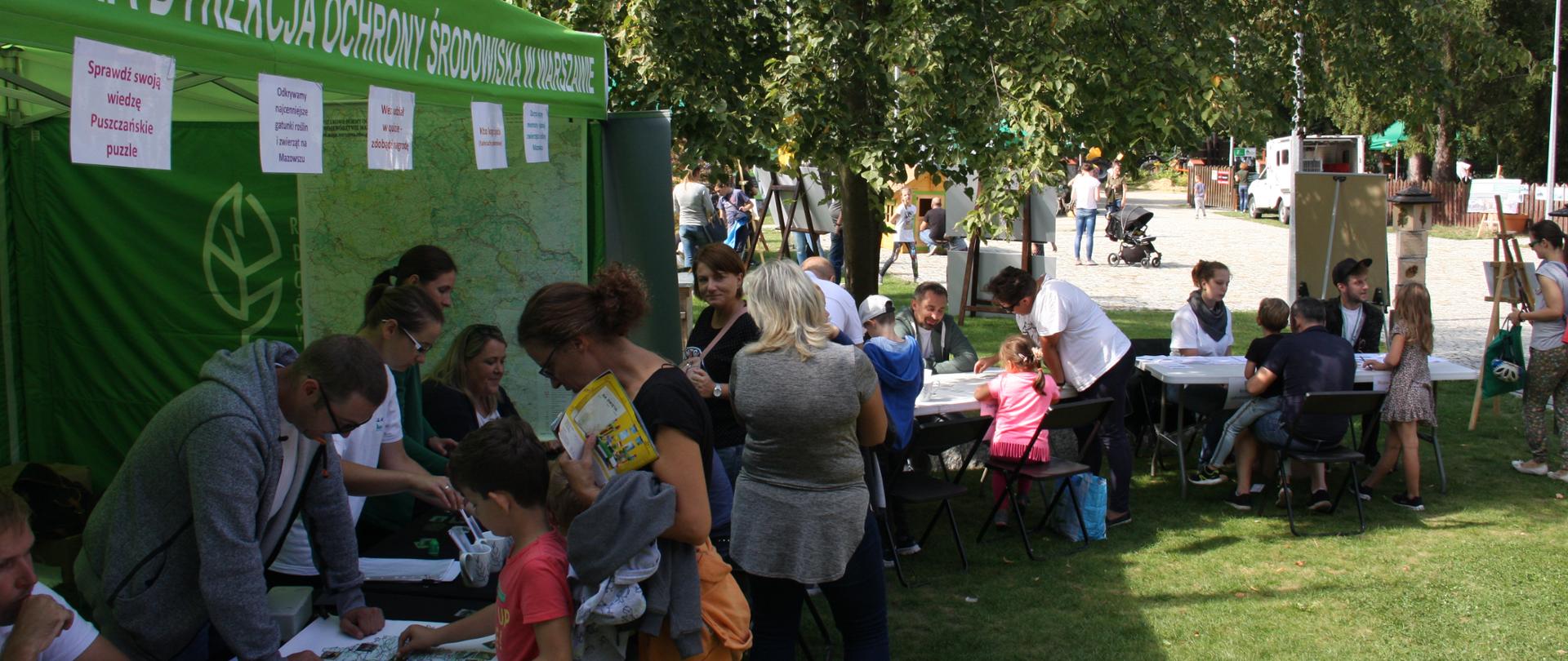 Po lewej stronie zielony namiot, przed nim stoją uczestnicy biorący udział w grach edukacyjnych, na drugim planie stoliki, przy których są prowadzone działania edukacyjne