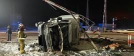 Wywrócony pojazd na przejeździe kolejowym. Strażacy wykonujący działania ratownicze. Pora nocna.