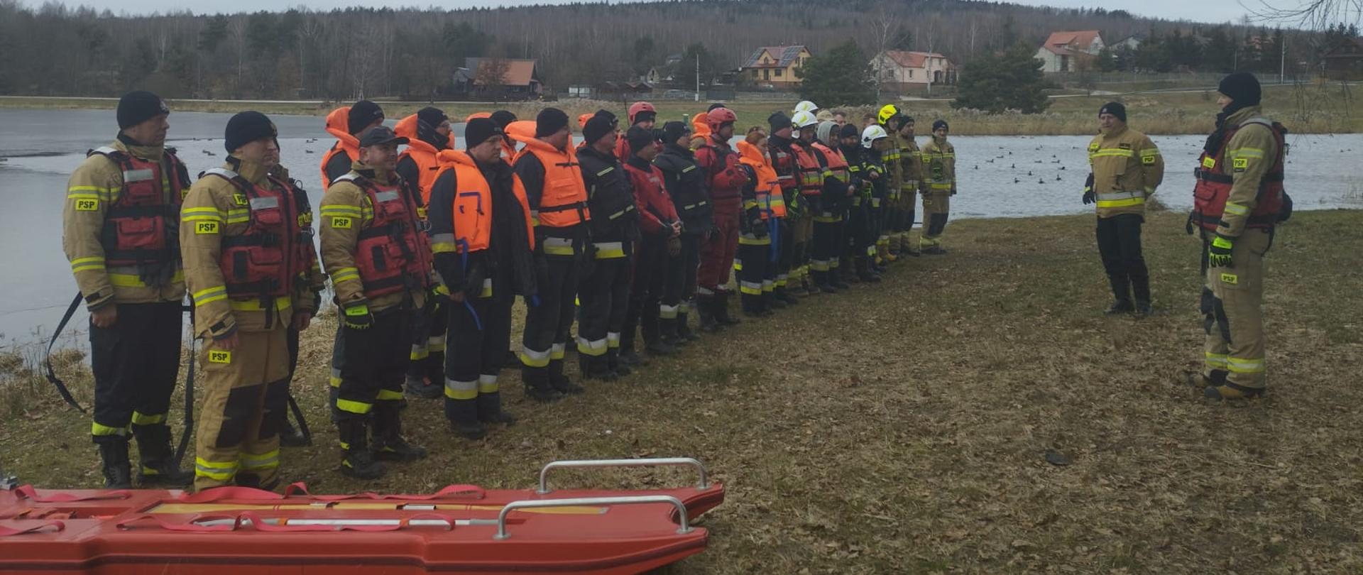 Zdjęcie przedstawia całą grupę ćwiczącą ratownictwo wodne nad zbiornikiem wodnym w Umrze podczas instruktażu teoretycznego do planowanych zajęć.