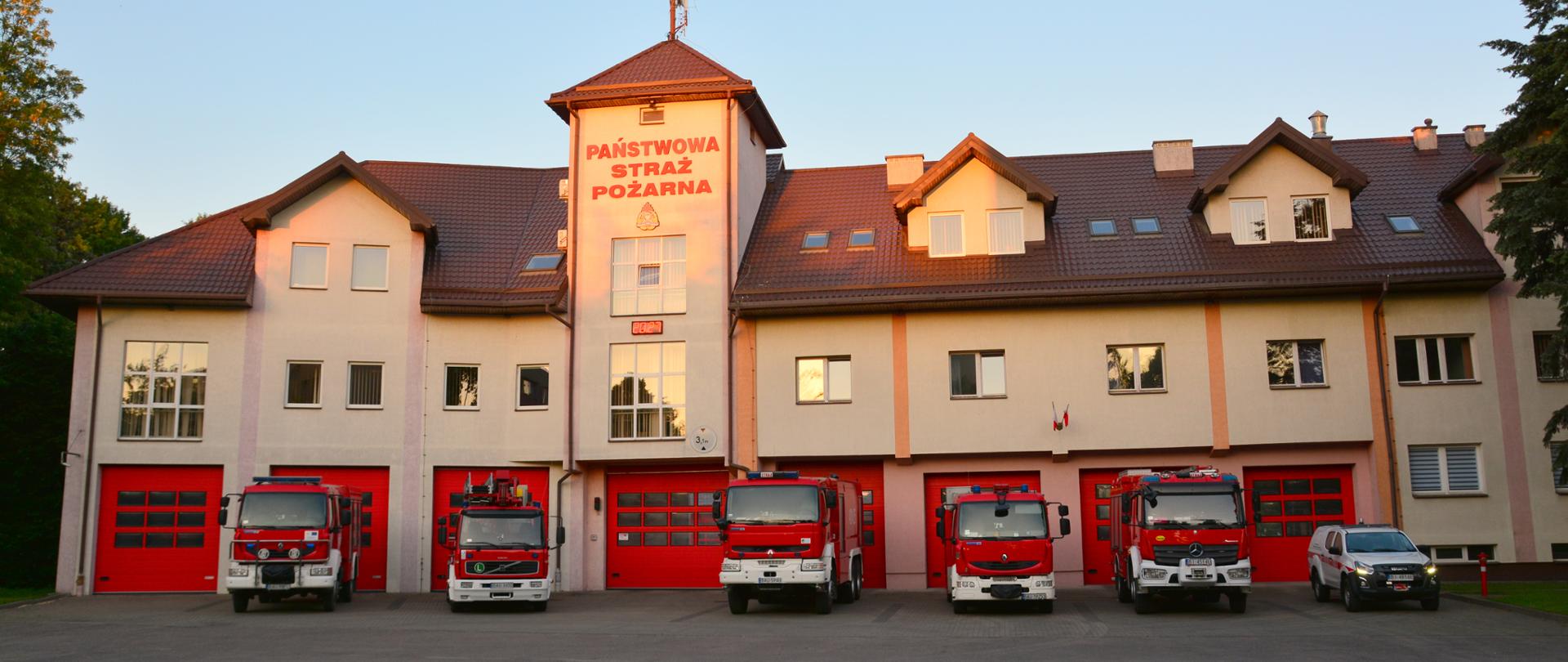 Na zdjęciu znajduje się budynek Komendy Powiatowej Państwowej Straży Pożarnej w Augustowie wraz z samochodami pożarniczymi.