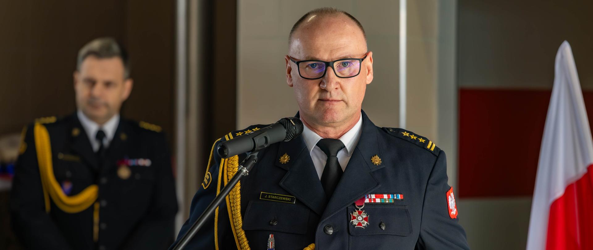 Strażak w galowym mundurze przemawia przy mównicy z mikrofonem. Na jego piersi widoczne są odznaczenia, a w tle stoi inny funkcjonariusz oraz polska flaga.