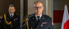 Strażak w galowym mundurze przemawia przy mównicy z mikrofonem. Na jego piersi widoczne są odznaczenia, a w tle stoi inny funkcjonariusz oraz polska flaga.