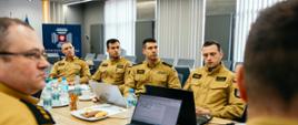 Grupa funkcjonariuszy Państwowej Straży Pożarnej w mundurach służbowych siedzi przy stole konferencyjnym z laptopami, dokumentami i napojami, uczestnicząc w spotkaniu w sali szkoleniowej Komendy Głównej PSP.”
